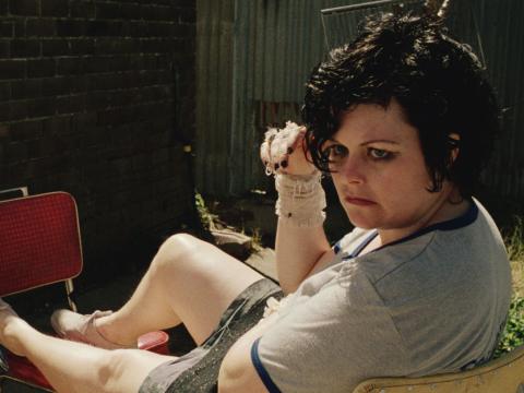 A woman sits outside with her legs outstretched on a chair. She looks back over her shoulder, scowling.