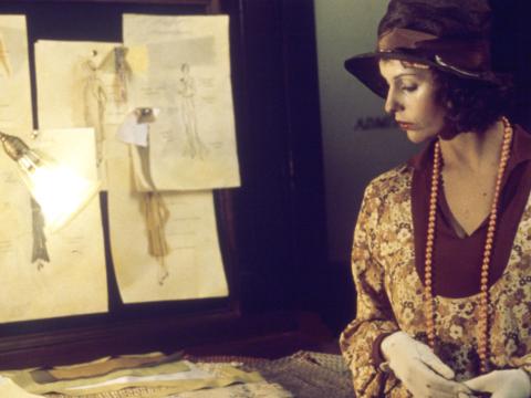 Film still of Helen Morse as Caddie looking at drawings of women's dress designs in a studio.