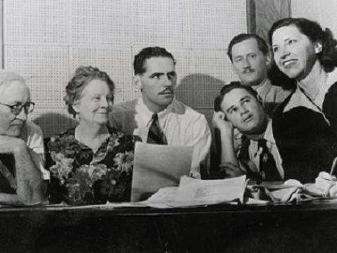 Grace Gibson with radio team gathered around a desk looking at scripts.