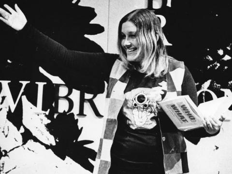 Lillian Roxon stands waving at the crowd holding a copy of a book in her other hand.