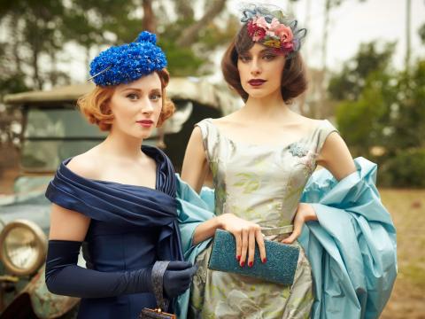 Actresses Amanda Woodhams and Hayley Magnus standing side by side looking at camera in 1950s style couture dresses and hats in a scene from the dressmaker. An old car can be seen in the background.