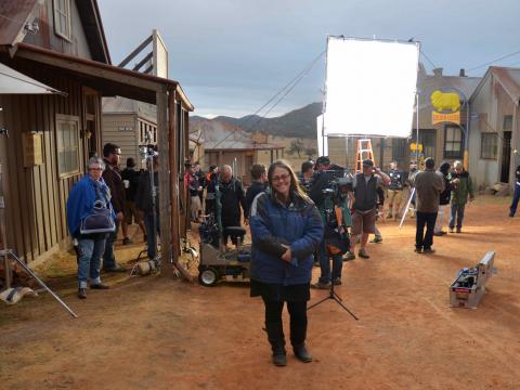 Director Jocelyn Moorhouse on the set of 'The Dressmaker', she is standing in a dusty red dirt road with old houses and buildings behind her and many members of the film crew working in the background.