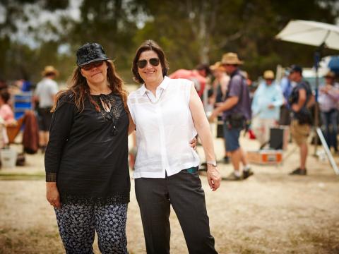 Director Jocelyn Moorhouse and Producer Sue Maslin on the set of 'The Dressmaker'. They are standing together smiling at the camera with a film crew working in the background. 