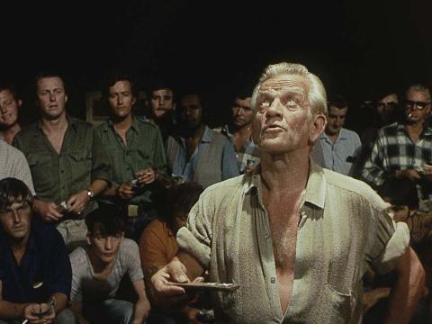 A man playing two-up in a dark room surrounded by a crowd of men