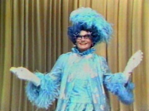 Dame Edna Everage poses for the camera, wearing a blue feathered gown and hat with white gloves.