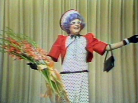 Dame Edna Everage brandishing her customary gladioli flowers in one hand with her arms outstretched in a welcoming way.