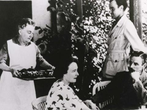 A family gathered with a older woman bringing a tray for tea.