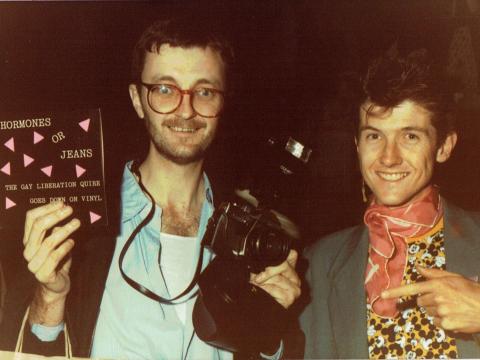 Two men standing next to each other posing for the camera. One is holding a camera in one hand and a vinyl record in the other. The record is entitled 'Hormones or Jeans: The Gay Liberation Quire Goes Down on Vinyl'.