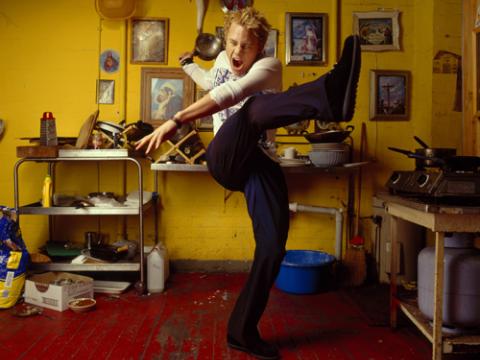 Actor Heath Ledger striking a dramatic pose with his arms out, one leg up in the air and his mouth open wide and eyes shut. He is in a yellow kitchen.