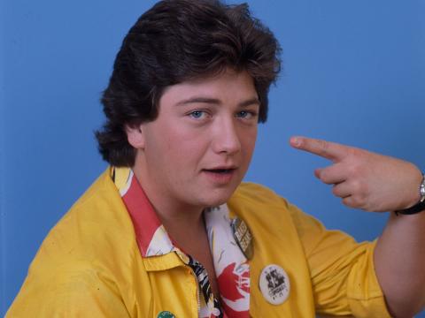 Jonathan Coleman wearing a yellow jacket, pointing a finger at his own face with bared teeth at camera.
