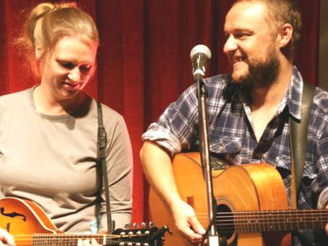 Jason and Chloe Roweth on stage performing against a red backdrop. Chloe playing banjo and Jason playing guitar.  