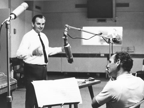 Two men in a recording studio. One seated playing a flute, the other standing and conducting.