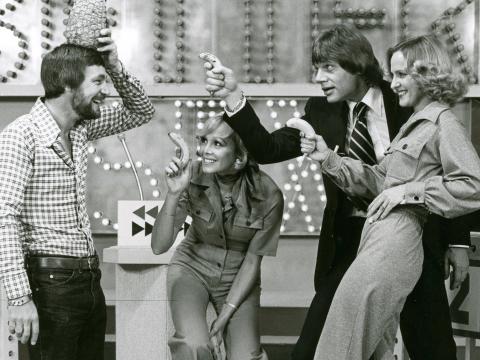Three people point at a man holding a pineapple on his head.
