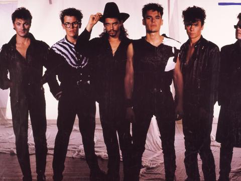 A promotional shot of INXS. The six band members are wearing black and standing in a studio, lead singer Michael Hutchence wearing a cowboy hat