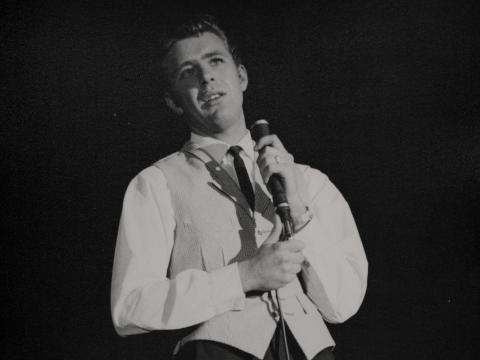 Lucky Starr wearing a shirt, tie and waistcoat singing into a microphone.