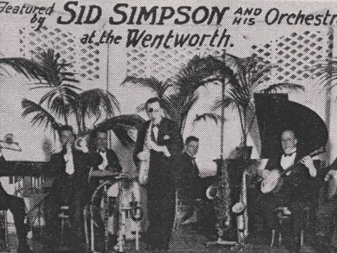Black and white newspaper image of an orchestra on stage surrounded by palm trees.