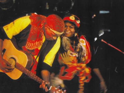 Archie Roach and Ruby Hunter performing on stage together. Archie is playing guitar and Ruby is smiling and they are leaning in towards each other. 