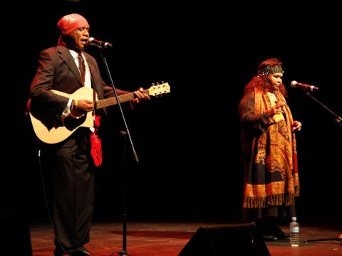 Archie Roach and Ruby Hunter on stage singing into microphones against a dark background. Archie is playing guitar.