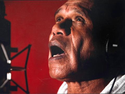 Close up of singer-songwriter Archie Roach singing into a microphone and looking upwards. He's wearing headphones and standing against a red background.