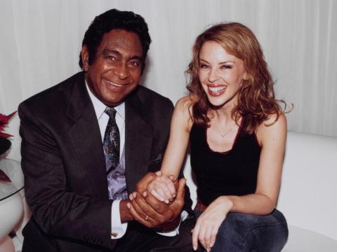 Jimmy Little and Kylie Minogue site on a sofa side by side. They are holding hands and smiling for the camera.