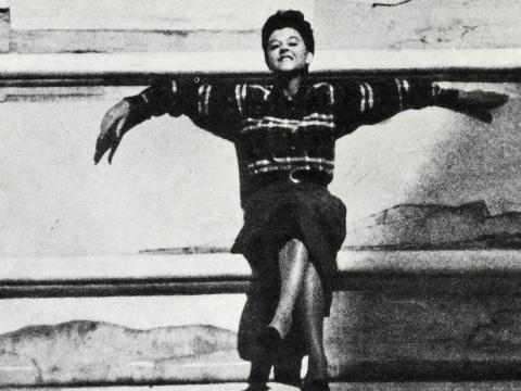 Composer Peggy Glanville-Hicks, seated with arms outstretched and legs crossed, smiling at camera. c. 1965
