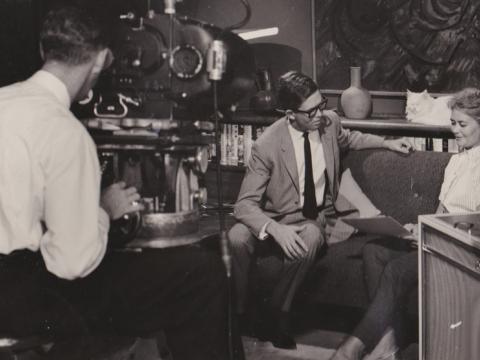 A man and a woman sit on a couch in a television studio talking with one another. Another man is filming them with a large camera.