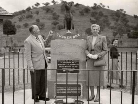 An older couple stand either side of a statue of a dog on a tucker box
