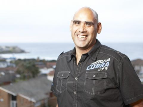 A smiling man in a black shirt smiles in front of a suburban view with the sea in the distance