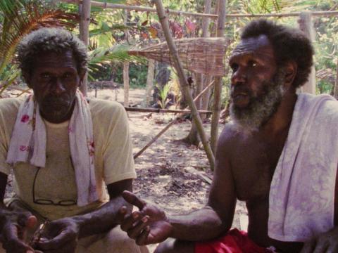 Eddie Mabo is seated on the ground, talking with another man.