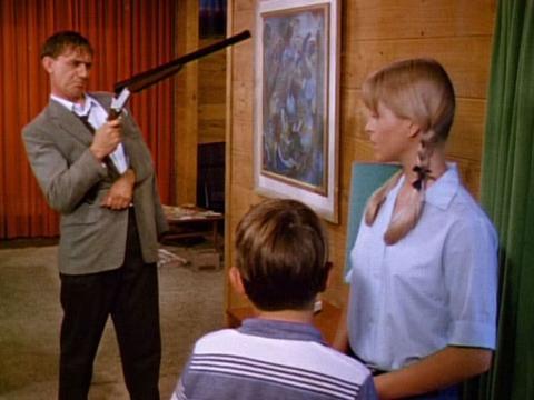 A man with his arm in a sling cocks a shotgun while a young woman and boy look on.