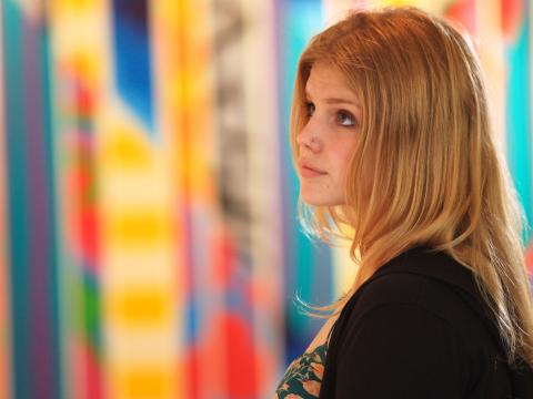 A young woman looking to her left is standing in front of colourful artwork in the background