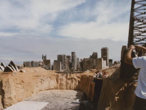 A crew member next to a model of the Sydney Harbour Bridge on the set of post-apocalyptic Sydney in some sand dunes. The Opera House and city skyline is also visible.