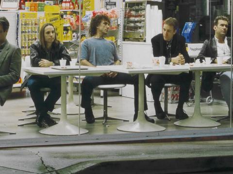 The five members of the band Cold Chisel seated at white plastic tables at a service station. From left to right: Don Walker, Phil Small, Ian Moss, Steve Prestwich and Jimmy Barnes.