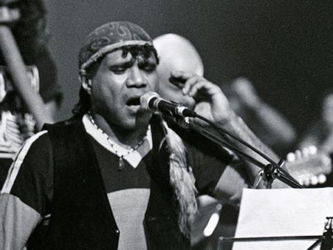 Close up of Indigenous singer Archie Roach singing into a microphone. Behind him on stage are other musicians in the background. 