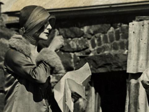 Director Paulette McDonagh outdoors directing a scene in a film. She is wearing a long coat and hat and looking through a camera.