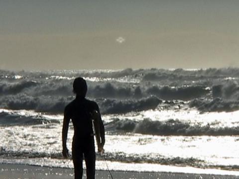 A silhouette of a man at the beach walking away from camera into the surf carrying a surfboard under one arm. 