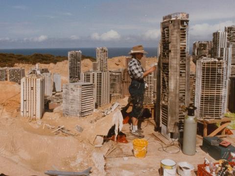 A crew member constructing a model set of post-apocalyptic Sydney in some sand dunes. The buildings are about the size of the person. The Opera House is visible.
