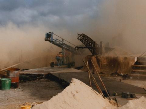 A crew member pushes a crane next to a model set of post-apocalyptic Sydney in some sand dunes. The buildings are about the size of the person. An arch of the destroyed Sydney Harbour Bridge and the Opera House is visible.
