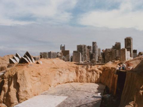 A crew member next to a model of the Sydney Harbour Bridge on the set of post-apocalyptic Sydney in some sand dunes. The Opera House and city skyline is also visible.