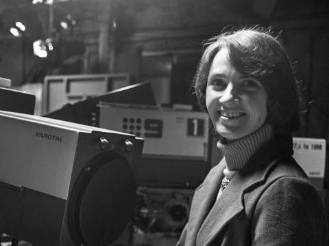 Woman standing behind some TV cameras in a studio, smiling directly at camera