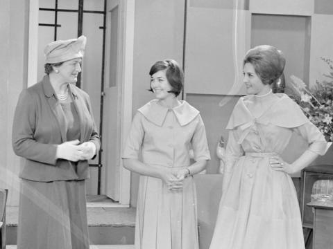 Three women standing in the middle of a television studio that is set to look like a suburban living room, c1960.