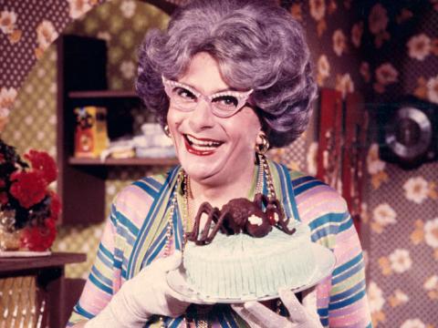 Dame Edna Everage is standing in a brightly coloured room. She has a mauve bouffant hairdo and her trademark winged glasses. She is holding an iced cake with a chocolate spider on top with gloved hands.