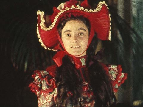 A young woman in a period floral dress with an ostentatious red, broad-brimmed hat