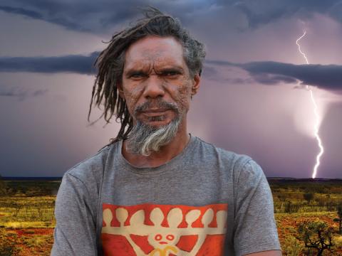 A bearded, middle-aged Aboriginal man in the outback stands staring at camera. In the distant background you can see lighting striking the ground.