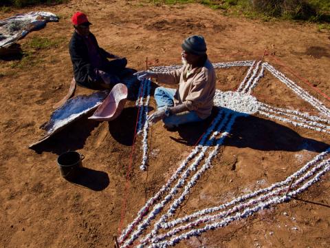 Two Aboriginal people sitting in the outback creating an image on the ground using white paint.