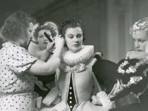 Vivien Leigh in costume on the set of Fire Over England has her hair adjusted by an assistant holding a comb as two other actors in costume look on