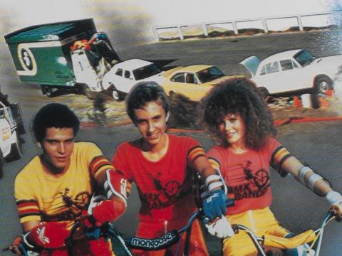 Three kids sitting on bicyles with some smoky wrecked cars in the background.