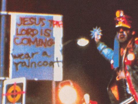 Menacing looking man about to throw something next to a graffiti sign that says 'Jesus the lord is coming. Wear a raincoat'.