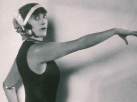 Black and white image of Annette Kellerman  wearing swimming costume and holding her arm out in front of her. She is looking at the camera.