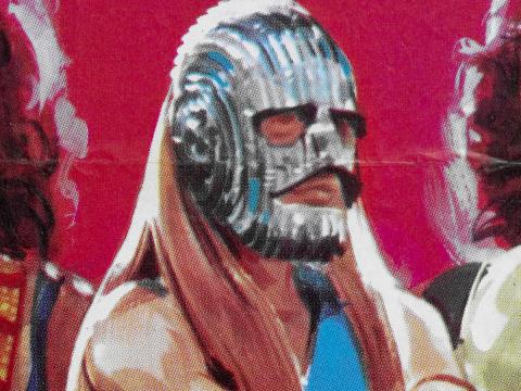 Close up head and shoulders poster art image of a muscled, long-haired man who has his face covered by a silver mask.